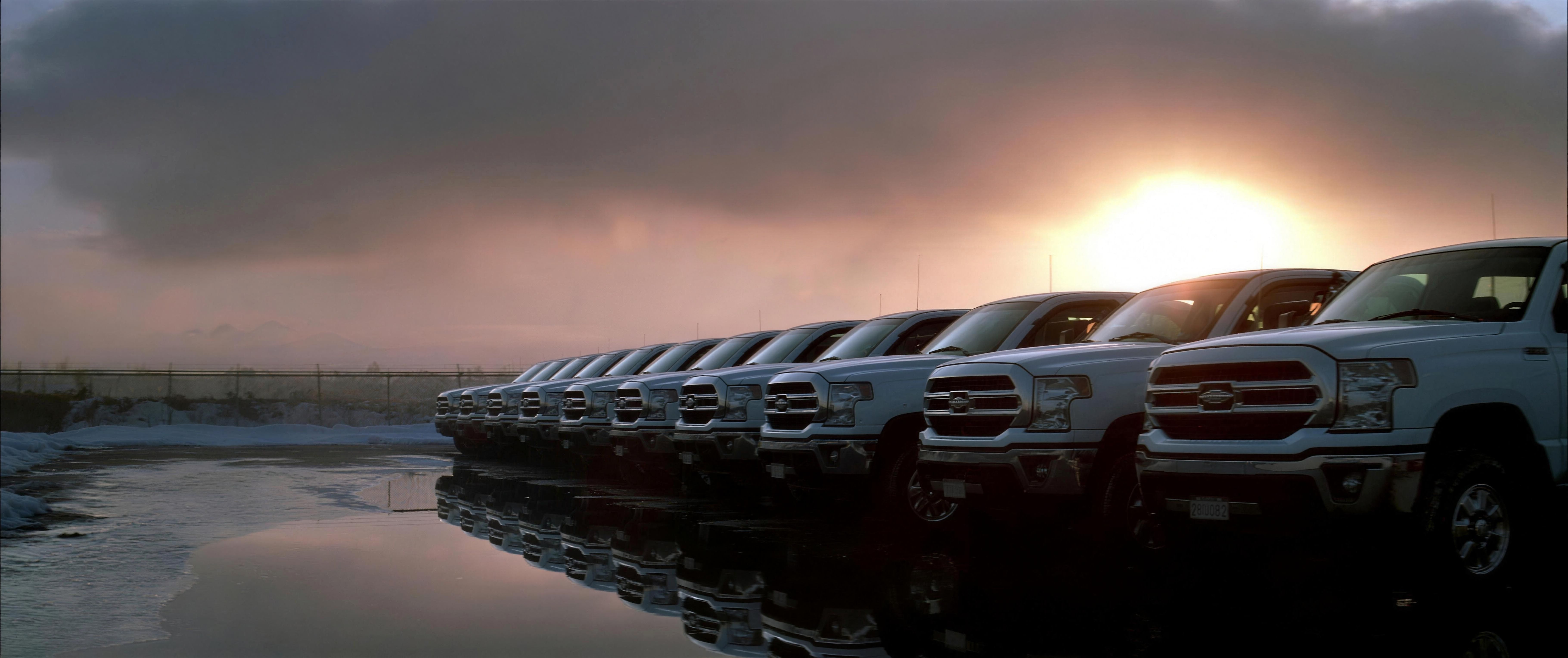 A uniform line of identical white GruMac pickup trucks parked nose-out along a flooded, mirror-still lot, a storm-lit winter sun breaking through dark clouds behind them