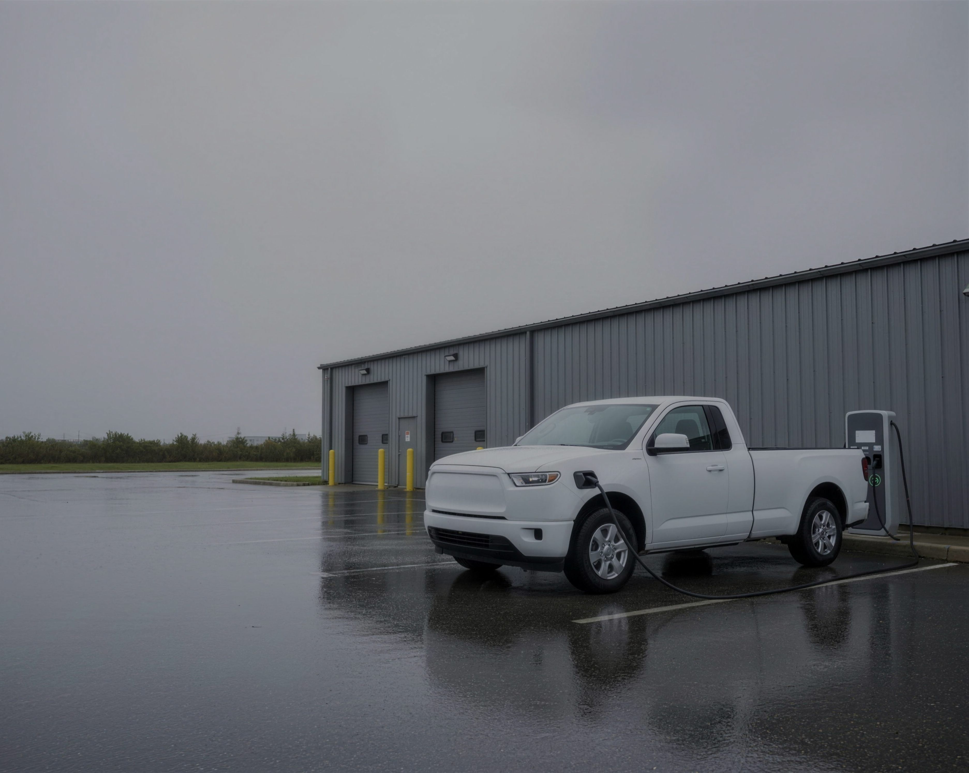 A white pickup truck plugged into an EV charging station on a wet depot apron under an overcast sky, with a grey metal-clad equipment building behind it