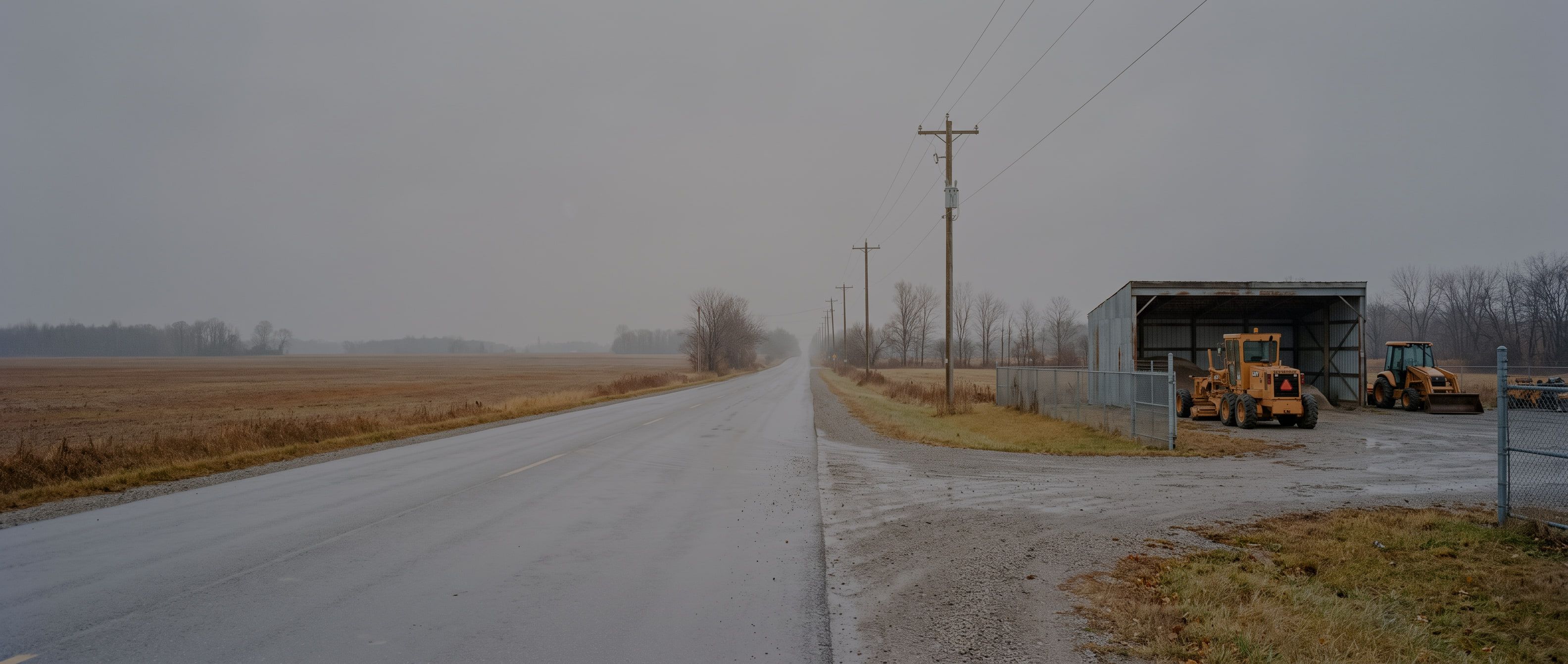 A wet rural Ontario concession road in late autumn running past a small municipal works yard, where a motor grader and a backhoe are parked beside an open-sided salt shed