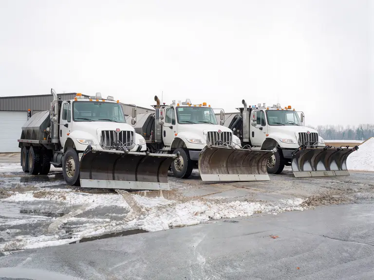 Three municipal snowplow trucks with front blades and salt spreader bodies parked in a row on a slushy works yard on an overcast winter day